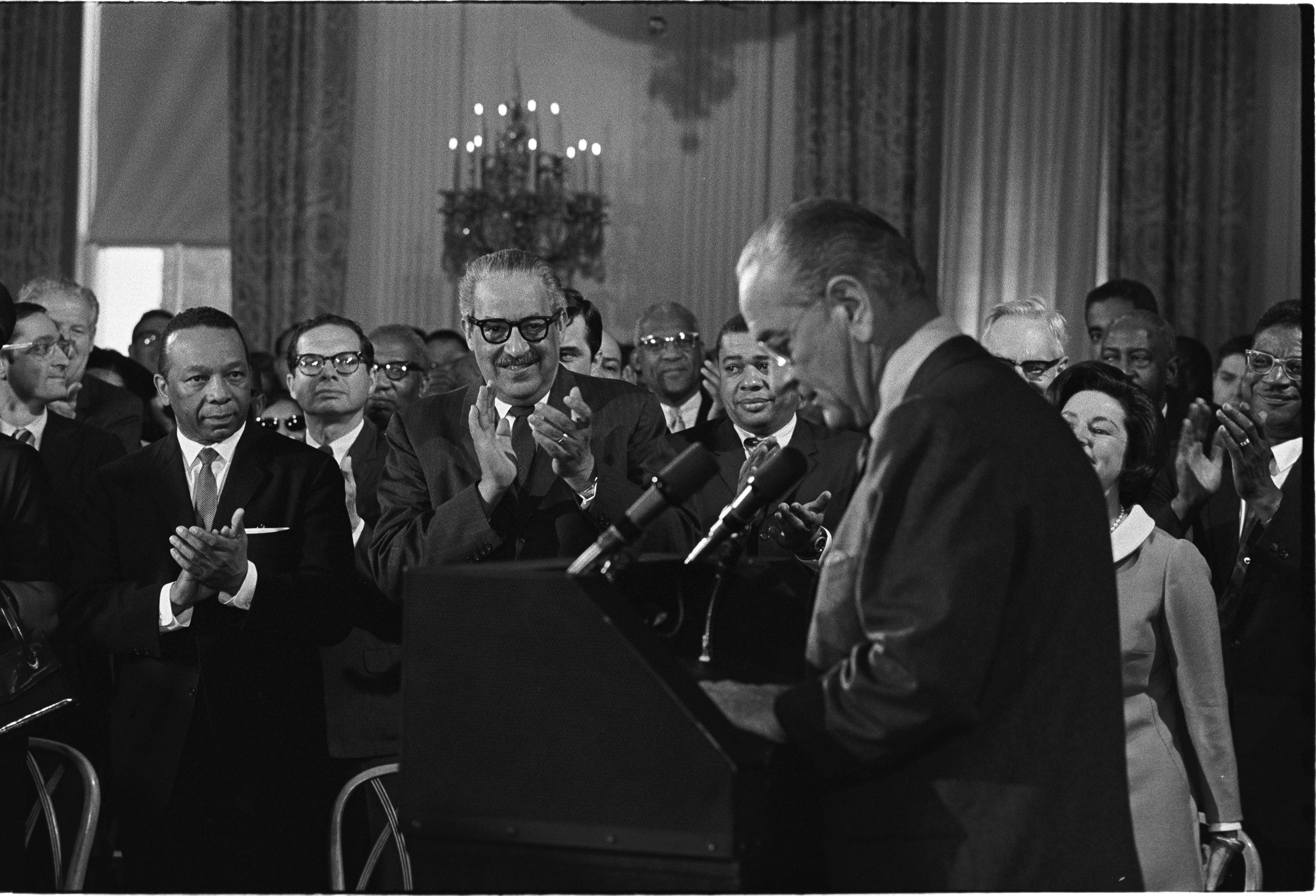 LBJ is in the foreground standing at a podium and wearing eyeglasses. His head is tilted forward as he reads his speech. Onlookers applaud.